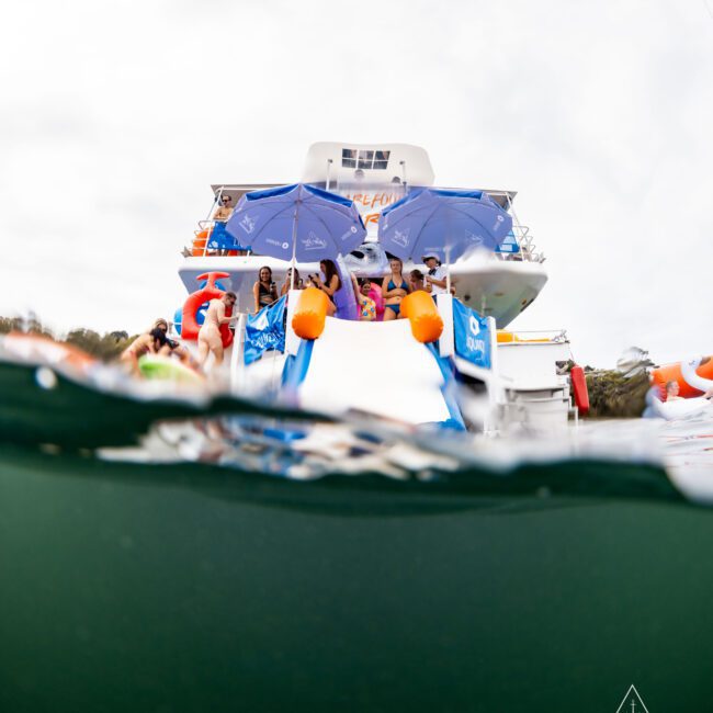 A group of people relaxing on a yacht with blue umbrellas and life jackets. The photo is taken from a low angle, partially underwater, capturing the vibrant scene above and the water below. Logo in the bottom right corner.