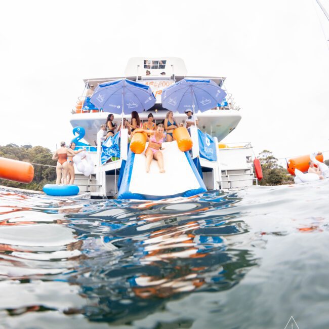 People are enjoying a yacht party, sitting and sliding on a water slide from the back of a boat into the sea. They're using orange and blue floaties. The boat is decorated with blue umbrellas, and the water is calm.