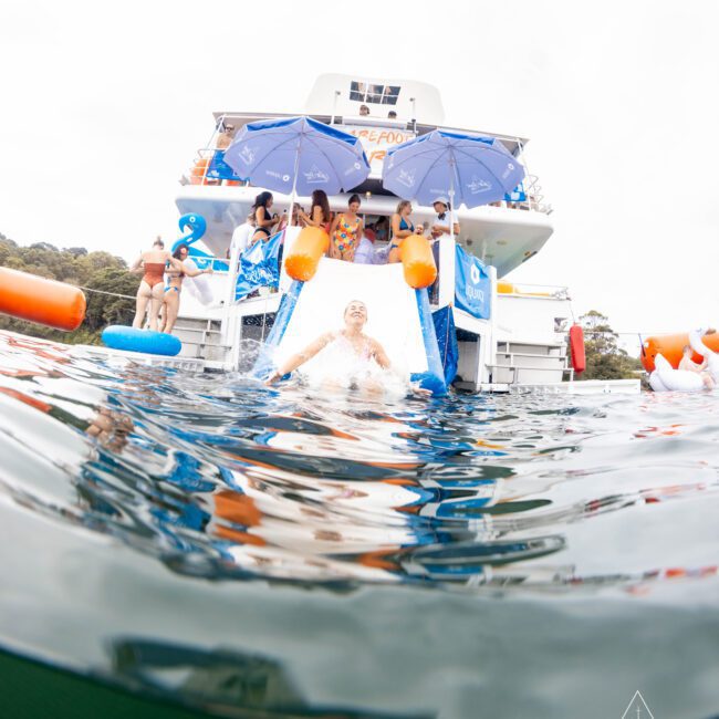 Person in swim trunks slides down a small water slide from the back of a boat into the water. People with umbrellas and inflatable toys are on the boat. The water surface and boat dominate the foreground and background.