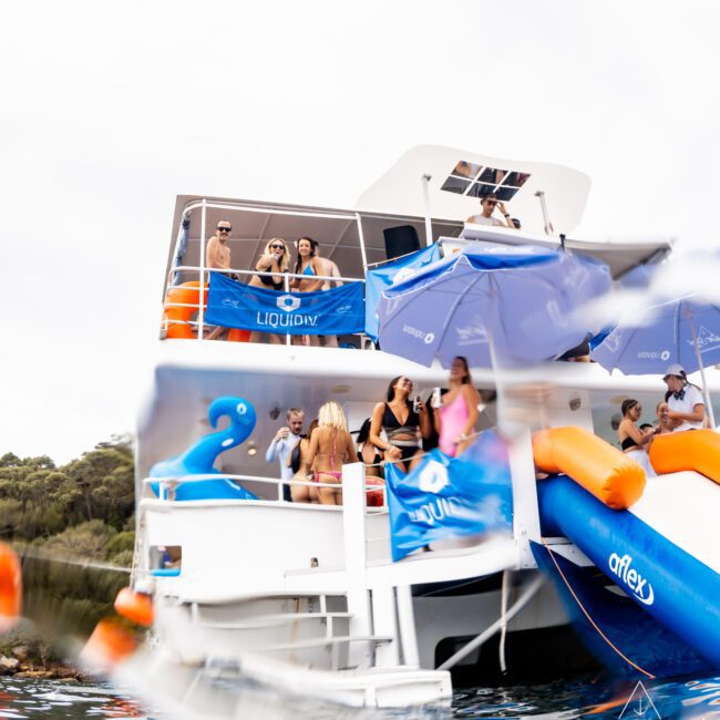 People on a white double-deck boat with blue banners, inflatables, and umbrellas. The image is partially submerged in water, showing a blue inflatable flamingo. Forested land is visible in the background.