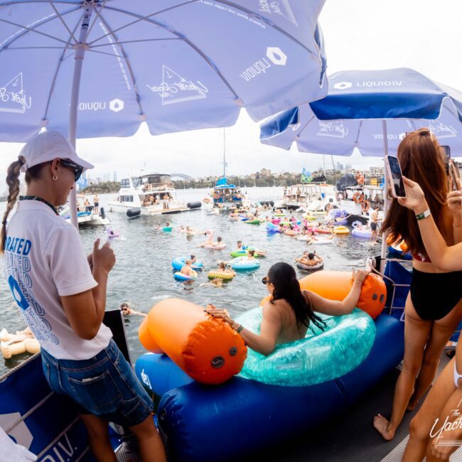 People on a boat take photos of a lively water party. Many are on colorful inflatables in the water. Boats are anchored nearby under a cloudy sky. Blue umbrellas with logo designs provide shade on the deck.