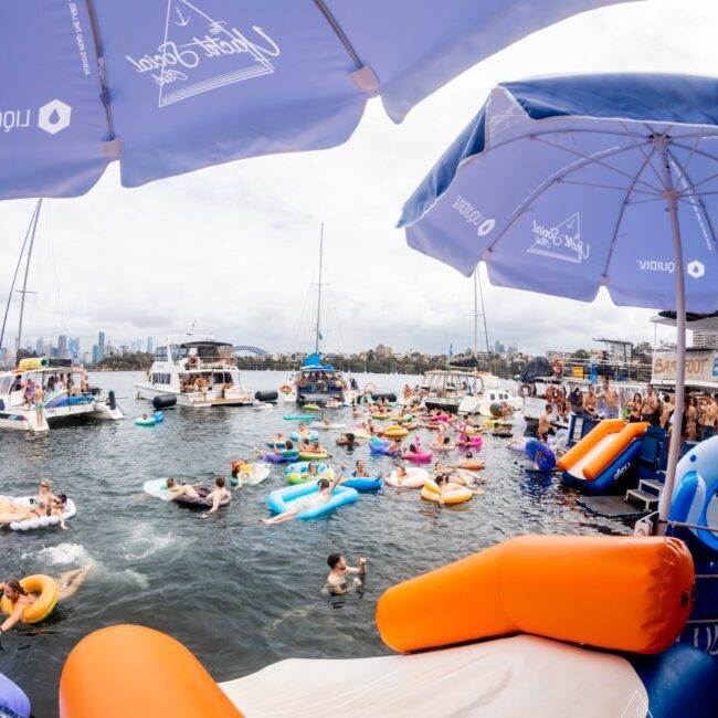 A lively scene at a waterfront event with people on colorful inflatables in the water. Boats and yachts are anchored nearby. The image is framed by large umbrellas, and there's an inflatable slide on one side. A city skyline is visible in the distance.
