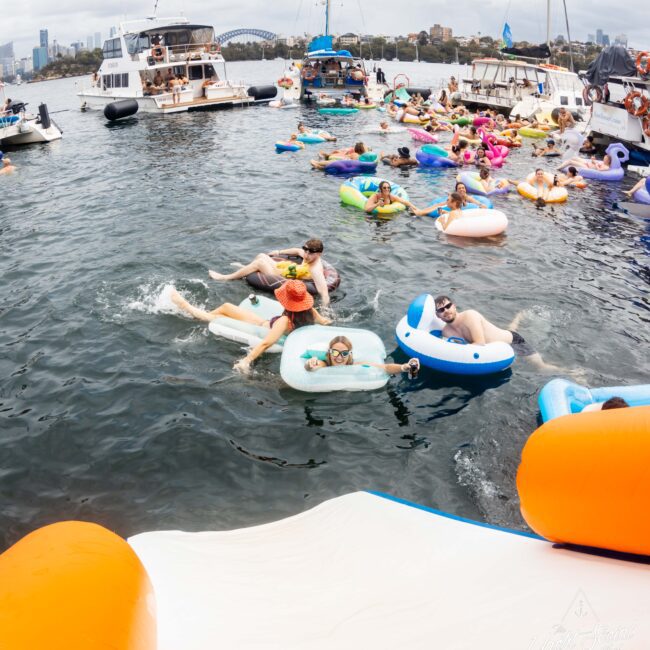 A group of people relax on inflatable floaties in a body of water surrounded by boats. The scene is lively and festive, with various colorful inflatables, under a cloudy sky with a city skyline in the background.