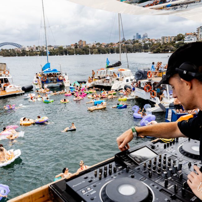 DJ performing on a yacht with a mixing console, overlooking a lively party on the water. People are floating on colorful inflatables, with boats and the Sydney Harbour Bridge in the background. A festive atmosphere under a cloudy sky.