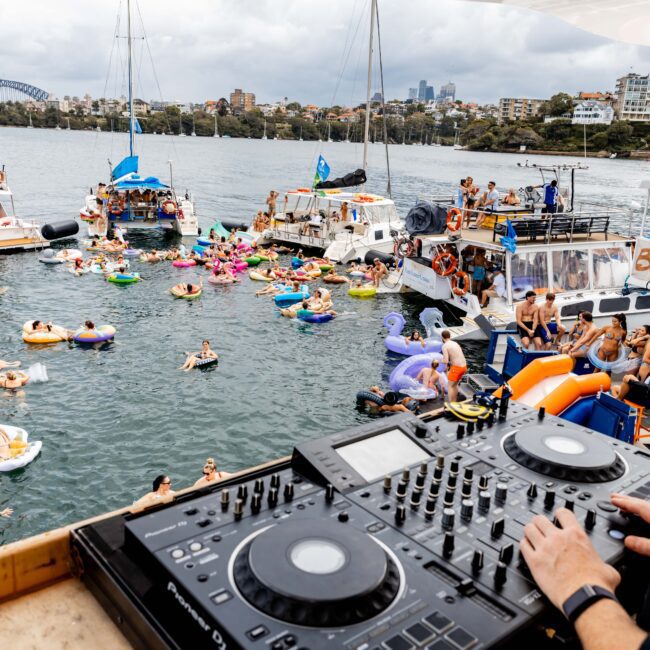 DJ playing music on a boat while people in colorful inflatables enjoy a floating party between anchored boats. The background shows a city skyline and a bridge under a cloudy sky.