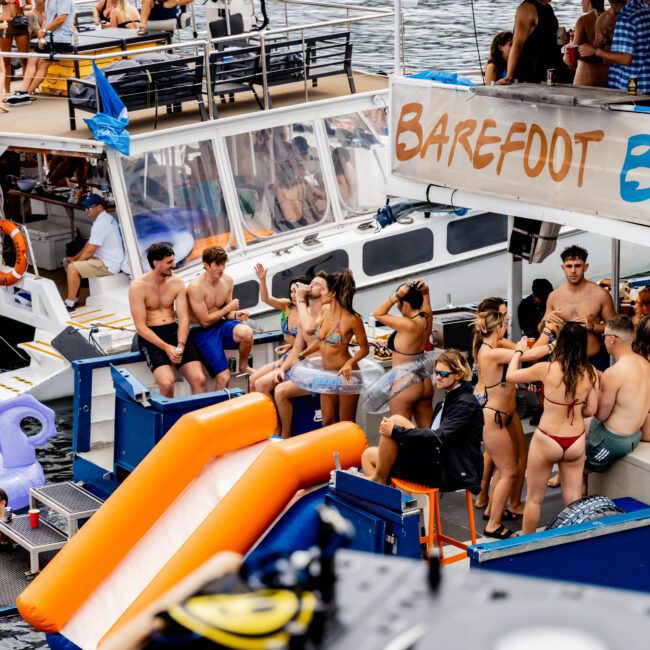 People enjoying a party on boats in a scenic water setting. Some are sitting on the deck, with others in swimsuits using a slide leading into the water. A "Barefoot" sign is visible. DJ equipment is in the foreground.
