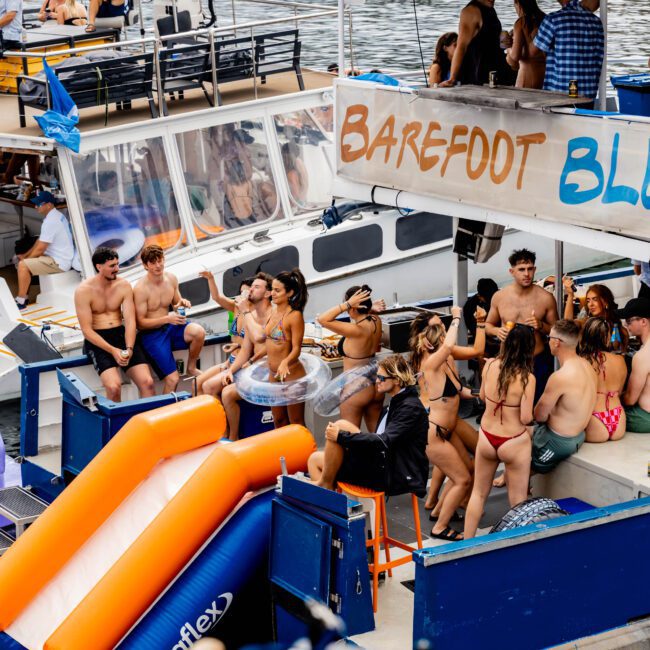 A group of people enjoying a sunny day on a boat named "Barefoot Blue". Some are sitting and chatting while others stand and dance. An inflatable slide is attached to the boat, and the water in the background is calm.
