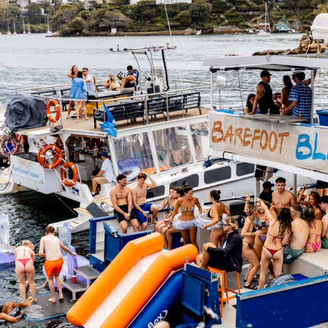 People enjoying a lively boat party on the water with colorful inflatables, music, and dancing. Multiple boats are anchored together, featuring a "Barefoot Blue" sign. Some guests are swimming, while others relax and socialize on the deck.