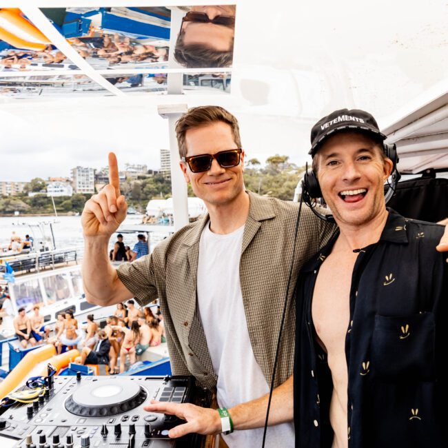 Two men are on a boat with DJ equipment. One is wearing sunglasses and a beige shirt, the other a black cap and shirt. A lively crowd and colorful inflatables are visible on boats in the background.