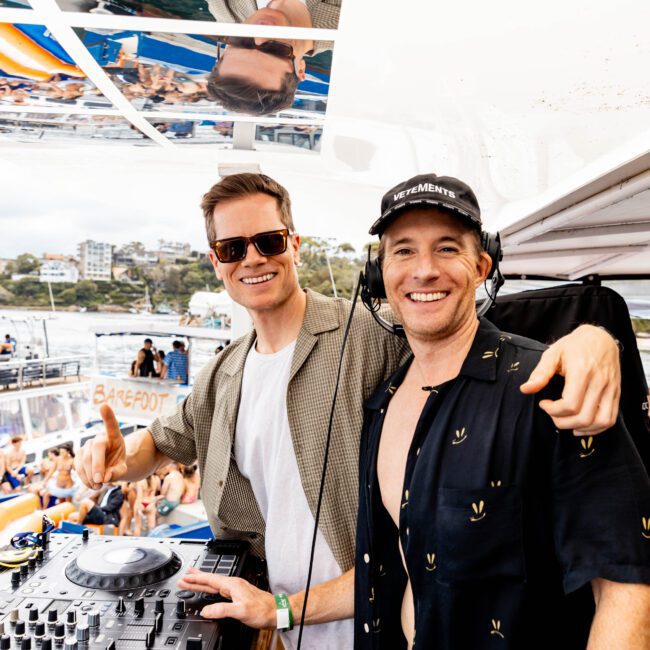 Two people smiling behind a DJ booth on a boat. One is wearing sunglasses and a beige shirt, and the other a black cap and shirt. A crowd enjoys the music, with water and a cityscape in the background.