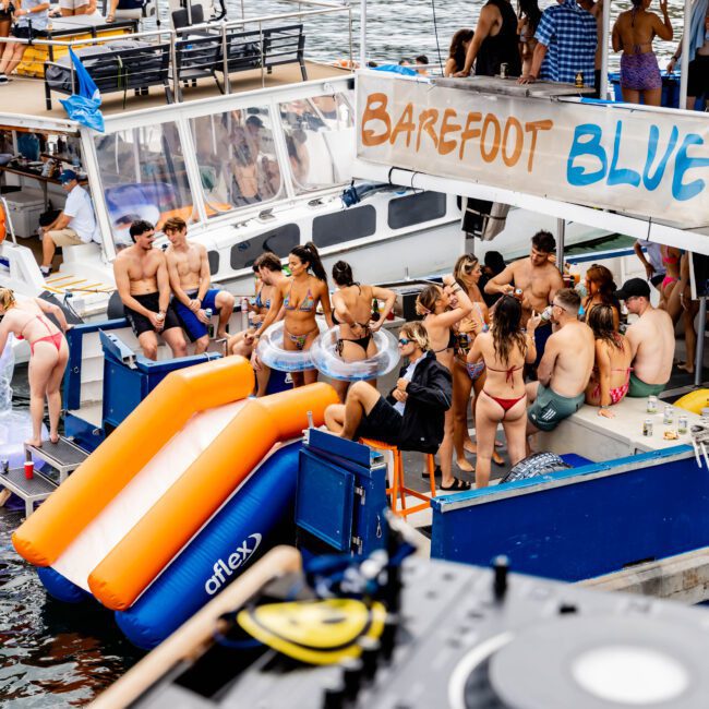 A lively scene at a boat party, featuring people in swimsuits socializing on and around two docked boats. A DJ setup is visible in the foreground. One boat displays a "Barefoot Blue" sign. Inflatable slides are attached to the boats.