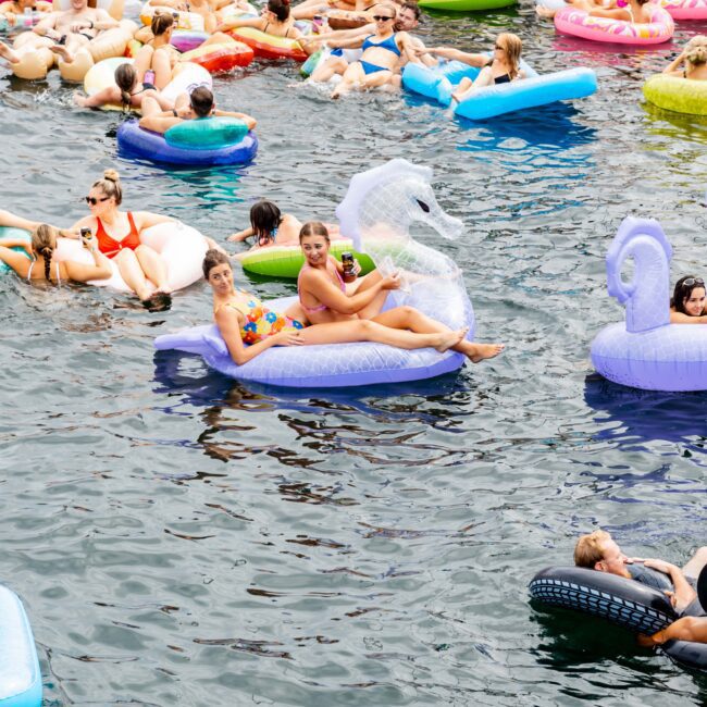 People float on colorful inflatable rafts in a crowded water area. The scene is lively, with many enjoying the water on a sunny day. Inflatable unicorns, swans, and other shapes are visible, along with some boats in the background.