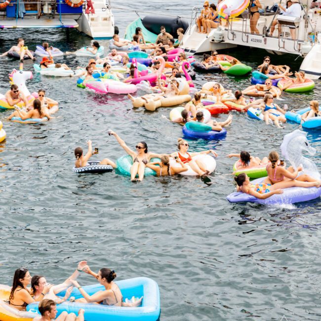 People are relaxing on inflatable floats in a body of water near boats. The scene is lively, with individuals interacting and enjoying a sunny day. Various float shapes and colors are visible, creating a festive atmosphere.