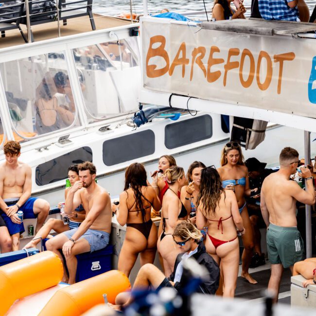 A lively group of people in swimwear enjoying a day on an anchored boat named "Barefoot." Some are lounging, while others are engaged in conversations and holding drinks. The water and part of another boat are visible in the background.
