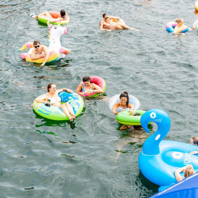 People on colorful inflatable floats, including swans and unicorns, enjoy a sunny day on the water. They are swimming and relaxing in the midst of a friendly gathering, creating a lively and fun atmosphere.