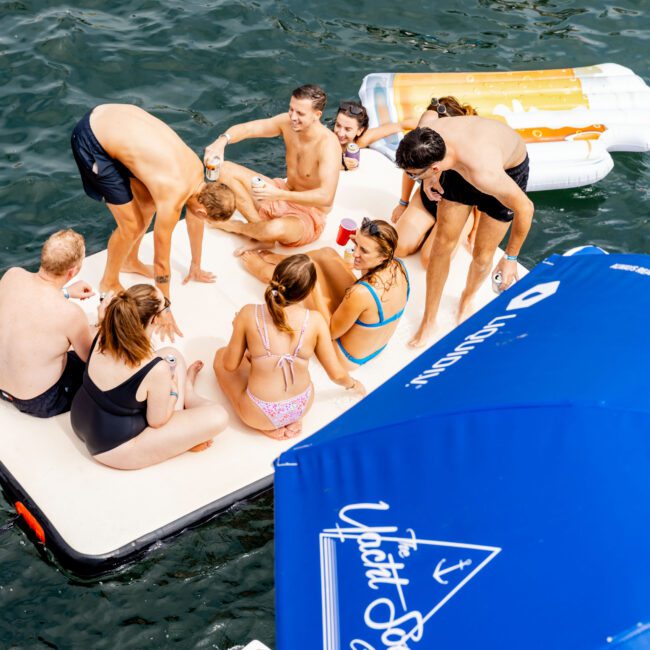 A group of people are gathered on a floating platform in the water, enjoying a sunny day. They are in swimwear and some are holding drinks. A blue umbrella with white text is partially visible in the foreground.