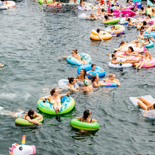 A lively scene of people floating on colorful inflatable rings in the water near a docked yacht. Inflatable shapes include a unicorn. The yacht is filled with more people enjoying the sunny day.