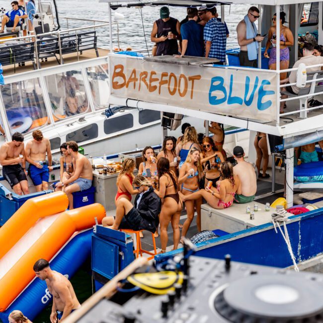 A group of people enjoying a party on a boat named "Barefoot Blue," featuring a DJ setup in the foreground, inflatable slides, and a sunny waterfront backdrop.