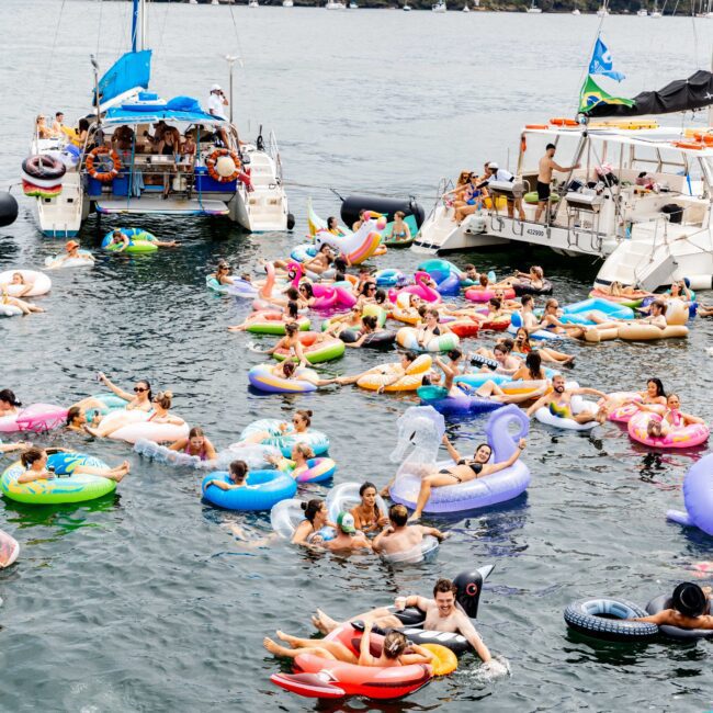 A lively scene of people enjoying a sunny day on a body of water, floating on colorful inflatable rings near two boats. The backdrop features a few sailboats and a hilly shore with buildings and trees under a cloudy sky.