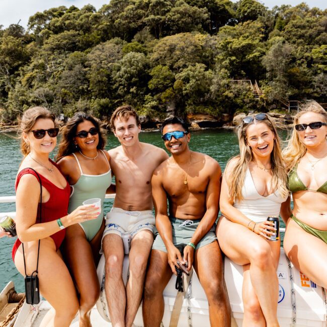 A group of five people in swimwear are sitting and smiling on a boat with tropical trees and water in the background. They appear relaxed and are enjoying the sunny day.