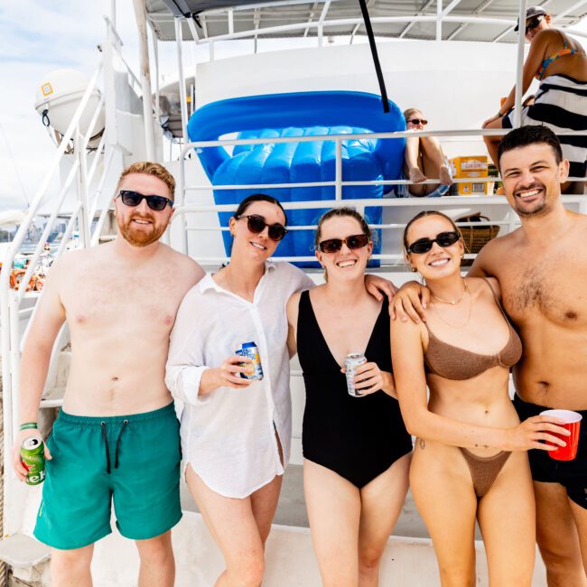 A group of five people is smiling on a yacht, wearing swimwear. The background shows a lifebuoy and more people relaxing. The sky is clear. Some hold drinks. The atmosphere is sunny and cheerful.