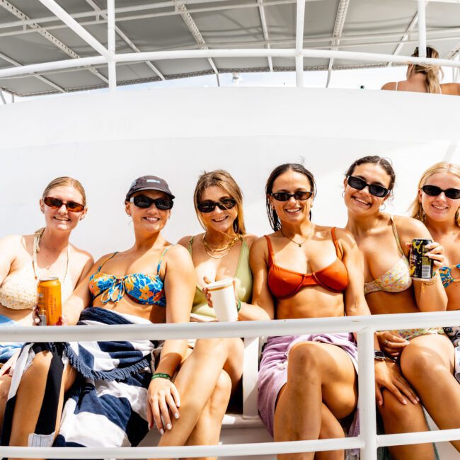 Six women are sitting together on a boat, smiling at the camera. They are wearing sunglasses and swimwear while holding cans and cups. The background shows a sunny environment with other people on the boat.