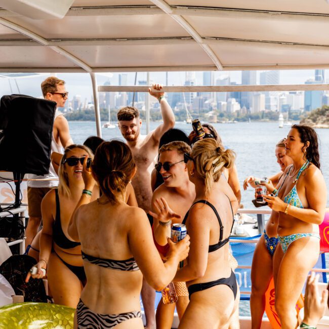 A group of people in swimwear are laughing and chatting on a boat. They are holding drinks and appear to be enjoying a sunny day. The background shows a city skyline and water.