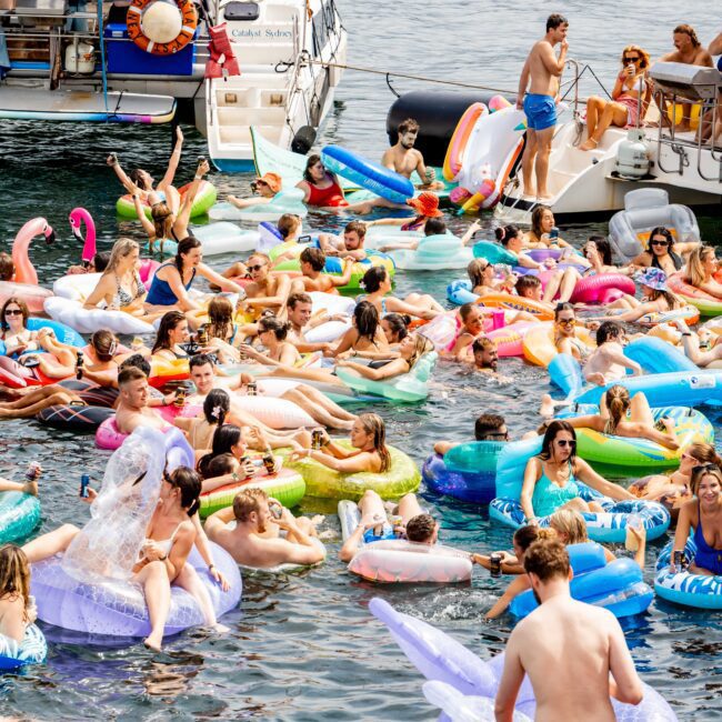 A crowded scene of people enjoying a sunny day on a lake, lounging on colorful inflatable floats. Two boats are docked nearby, with additional people on board. Many are in swimwear, splashing and soaking up the sun.