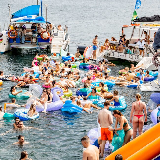 A lively scene of people enjoying a summer day on the water with various colorful inflatables. Yachts are anchored nearby, and the Sydney Harbour Bridge is visible in the background. The atmosphere is festive and fun.