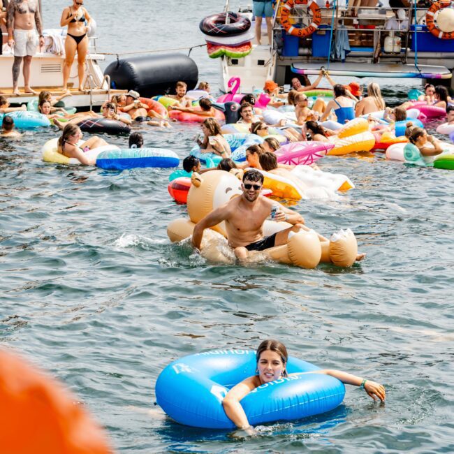 People enjoy a sunny day on a river, floating on colorful inflatables near boats. Inflatables include animals and geometric shapes. There are men and women in swimwear, some lounging on boats, with a bridge visible in the background.