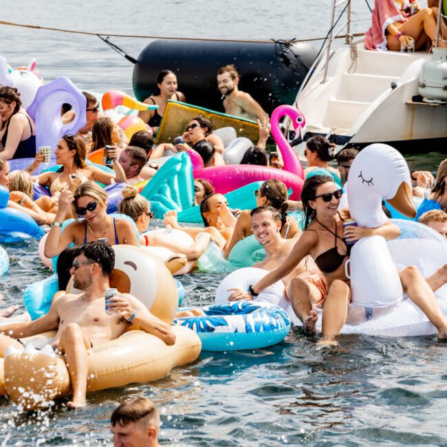 A lively scene on the water with people relaxing on colorful inflatable floats, including flamingos and unicorns. A boat is visible in the background, and several individuals are enjoying the sunny day, holding drinks and socializing.