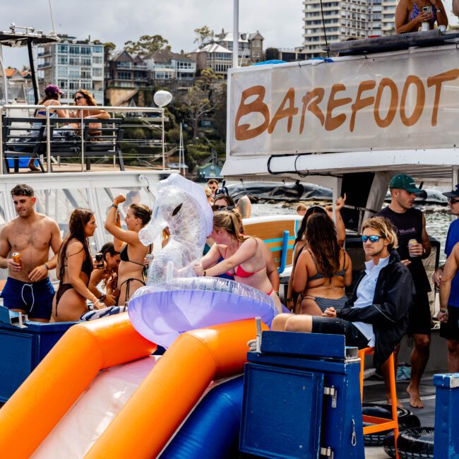 People enjoying a boat party. A group is gathered around a water slide, and a man wearing a suit jacket sits nearby. Others socialize with drinks in hand. The boat has "Barefoot" written on a banner, with city buildings visible in the background.