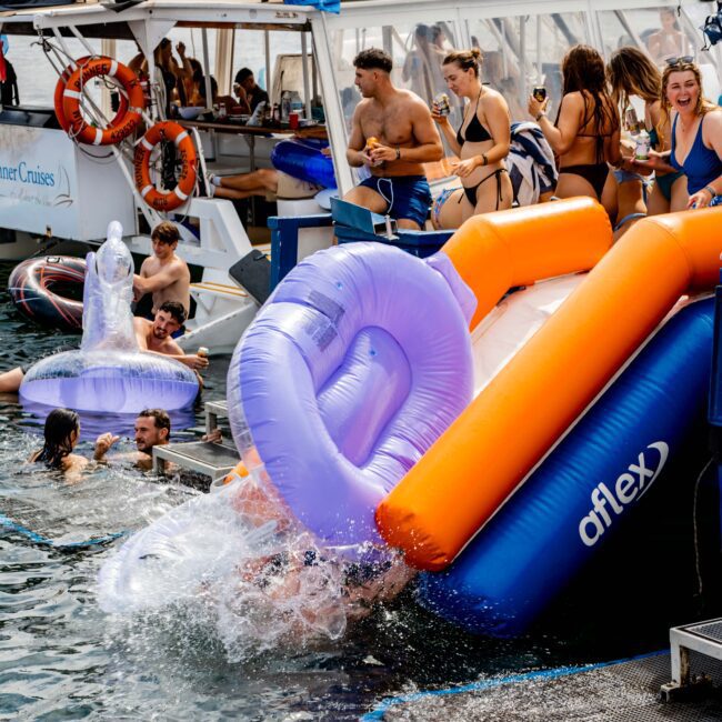 People enjoying a boat party, with some swimming and others using a large inflatable slide attached to the boat. Inflatable tubes are seen, and life rings are hung on the boat. The background shows additional boats and buildings.