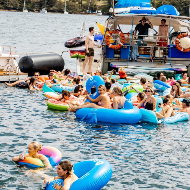 People enjoying a sunny day on the water, floating on colorful inflatables near boats. In the background, a bridge and buildings are visible. The scene is lively and festive, with various inflatable designs like an octopus and a donut.