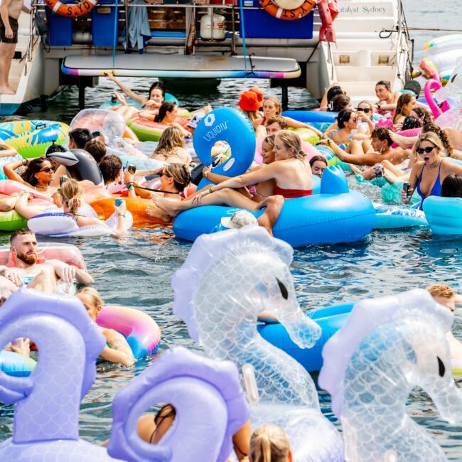A lively group enjoys a summer day on the water, surrounded by colorful inflatables, including unicorns and flamingos. A boat anchored nearby adds to the festive atmosphere, while people lounge and socialize on their floats.