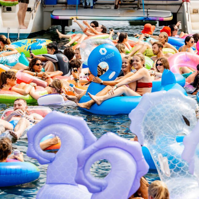 A crowded pool with people relaxing on colorful inflatable floats. Various shapes like flamingos and swans are visible. A boat is in the background with people sitting on the deck. Everyone is enjoying a sunny day on the water.