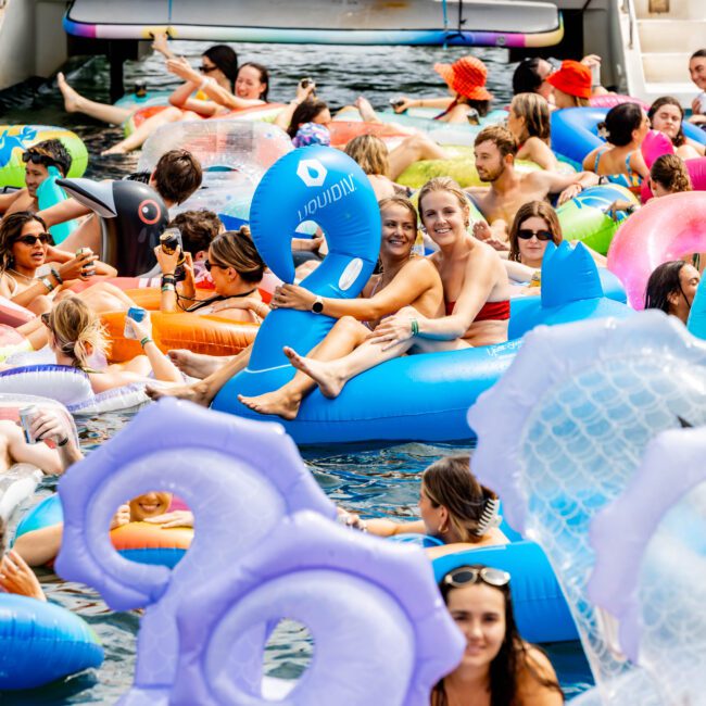 A lively pool party on a sunny day, featuring numerous people relaxing and socializing on colorful inflatable floats. The floats include various shapes, with a boat in the background and a clear, vibrant atmosphere.
