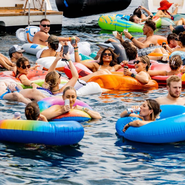 A lively group of people enjoying a sunny day on the water, floating on colorful inflatable rafts and rings near boats. They are socializing, smiling, and holding drinks, surrounded by a festive atmosphere.
