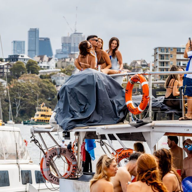 A lively boat party with people in swimwear enjoying the day. Two women pose for a photo on the upper deck, with city buildings in the background. The sky is overcast, adding a lively yet relaxed atmosphere to the gathering.