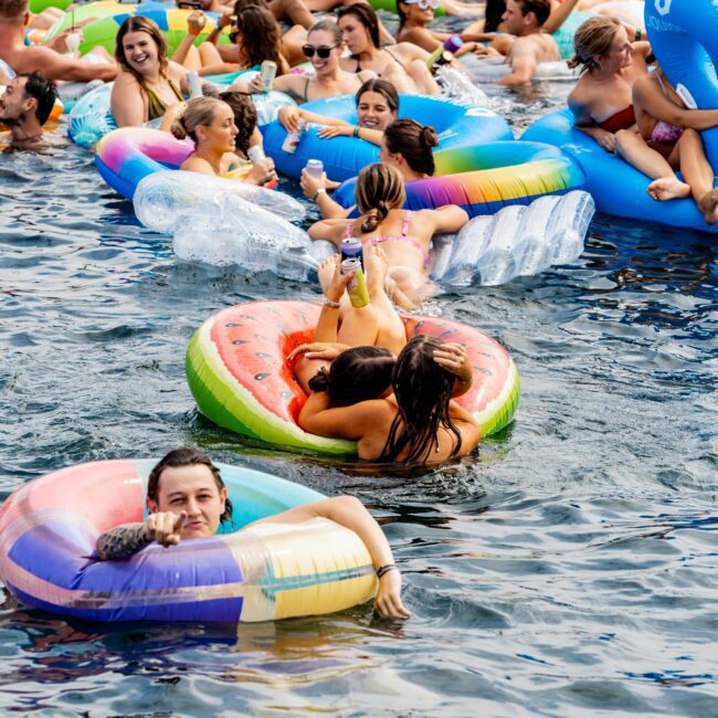 A lively pool party scene with numerous people relaxing on colorful inflatable pool floats in the water, including a watermelon slice and a rainbow design. Some people are drinking and socializing, while others enjoy the sunny day.