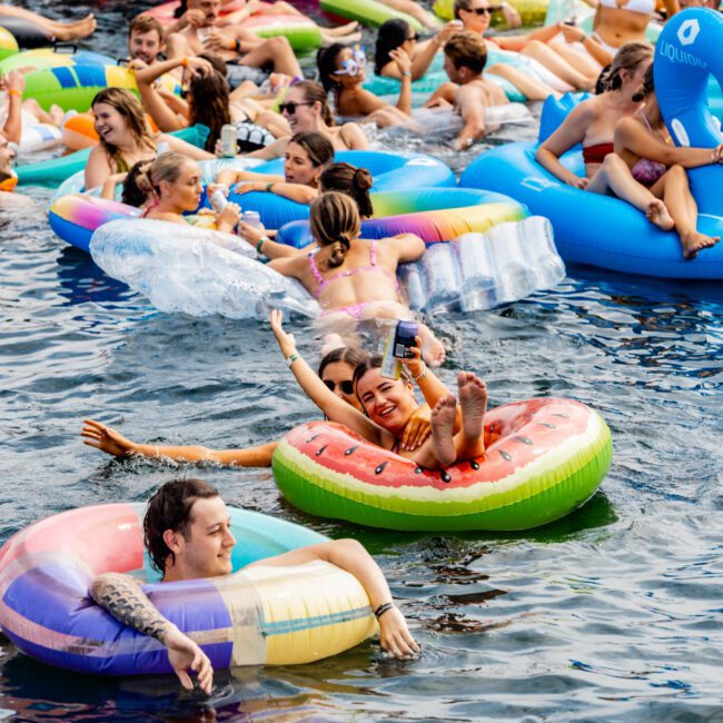 A lively group of people enjoying a pool party in the water with various colorful inflatables, including donuts and a watermelon slice. They are relaxing and socializing under a sunny sky, with a boat in the background.