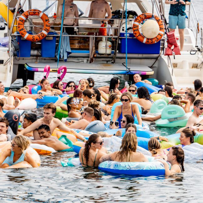 A crowded gathering of people in a lake, enjoying the water on various colorful inflatables, near a boat. The scene is lively, with smiling individuals under sunny weather and lush green trees in the background.