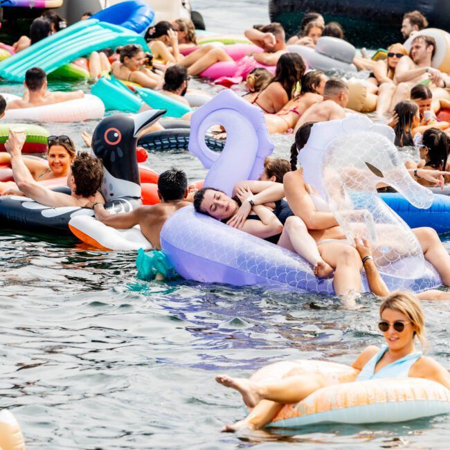 People are lounging on colorful pool inflatables, including swans and a duck, in a crowded lake setting. Some are socializing, while others relax and enjoy the sunny weather. A boat is visible in the background.