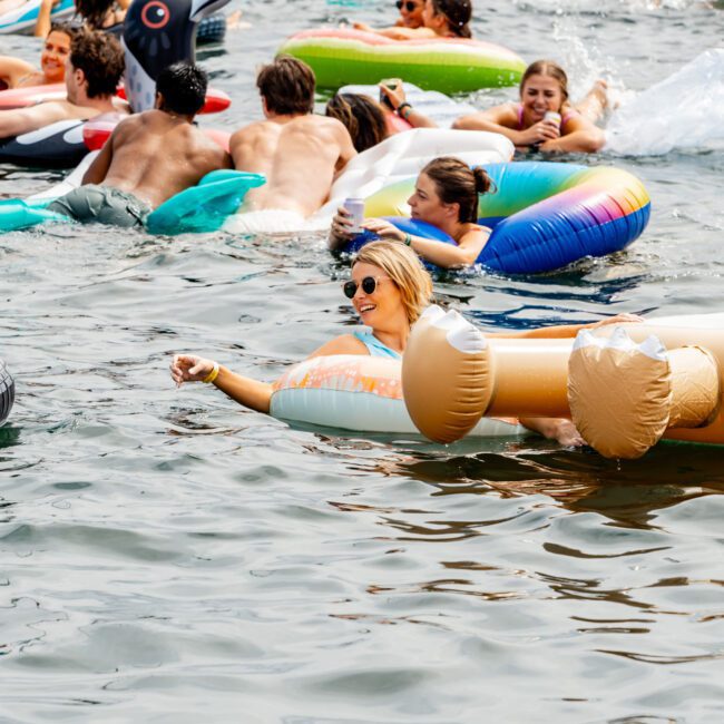 A group of people lounging on inflatable floats in a large body of water. Some floats are shaped like animals, while others are plain. One person in sunglasses is prominently floating on a large circular float. The mood is relaxed and fun.