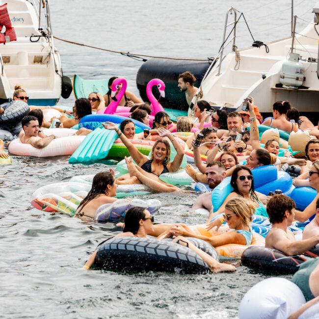A lively gathering of people in a water body using various inflatable floats, including flamingos, a tire, and a pineapple. Boats are anchored nearby, and the atmosphere is festive, with individuals smiling and enjoying the water.