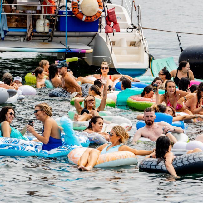 A group of people relax on brightly colored inflatable floats in a body of water. They are gathered around a boat, and some hold drinks, enjoying a sunny day. There are trees and a sailboat in the distant background.