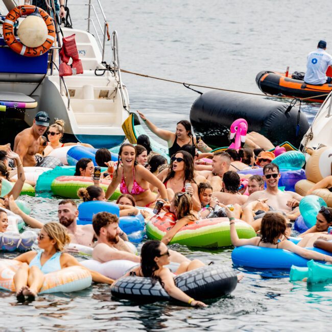 A group of people on colorful inflatable rafts enjoy a party on the water near several boats. The scene is lively and festive, with some attendees wearing swimsuits and others interacting with large floats. The background features a calm body of water.