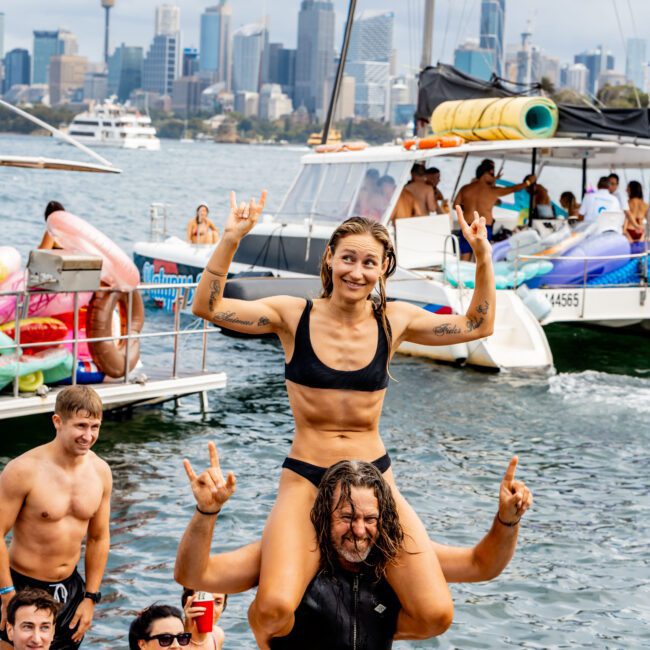 A woman in a black bikini sits on a man's shoulders, both smiling and making rock and roll hand signs. They are surrounded by others in swimwear enjoying the water, with boats and a city skyline in the background. The atmosphere is lively and festive.