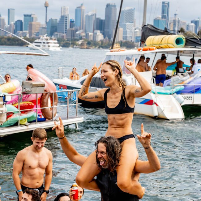 A man holds a woman on his shoulders in a lively boat party setting. Both are smiling and making hand signs, with the city skyline in the background. Others around them are enjoying the water.