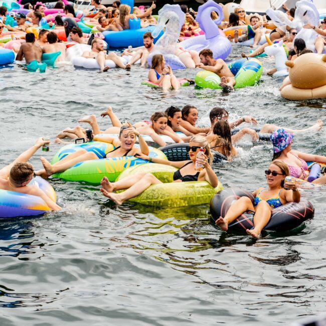 A lively gathering of people floating on colorful inflatable rafts in a body of water near a boat. Participants are enjoying a sunny day, some holding drinks. Various floats include swans and flamingos. The atmosphere is festive and social.